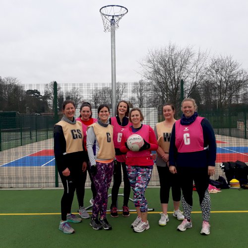 A group of women standing in front of a netball hoop with a netball, all smiling and wearing team bibs