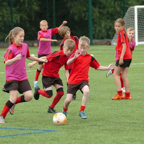 A group of boys and girls dressed in red playing football on an astroturf pitch